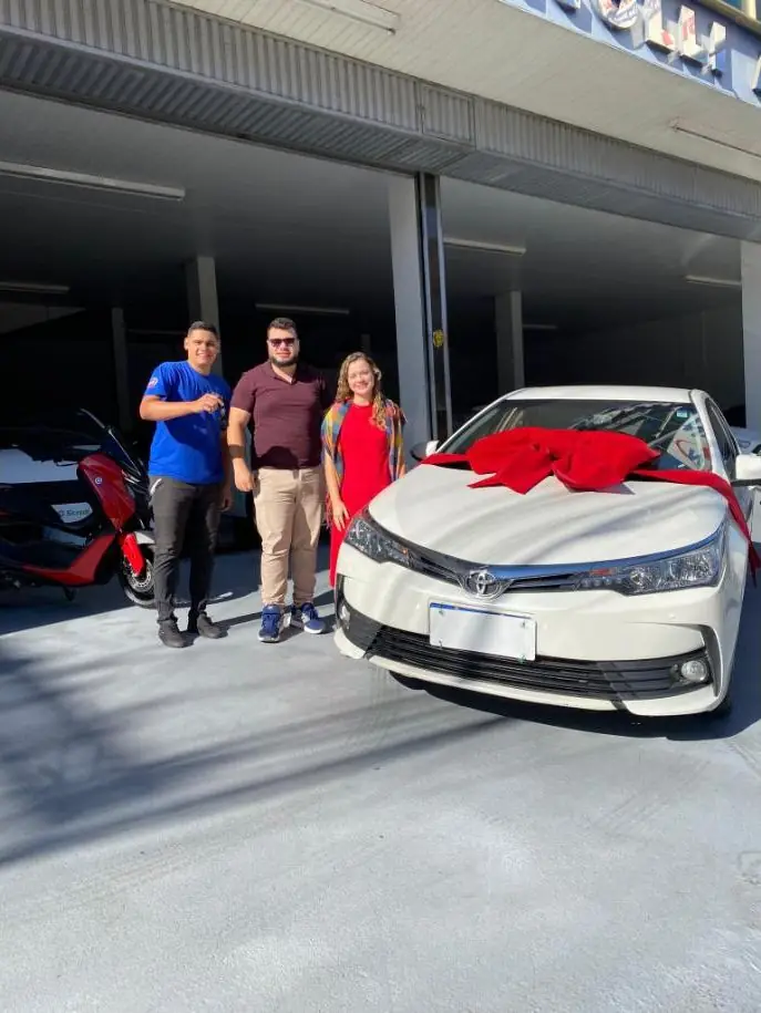 Lucas com um casal ao lado de um Toyota Corolla branco com laço